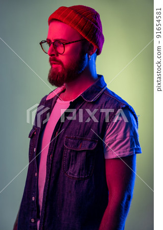 Portrait of confident bearded male looking to side space with serious attentive face, wearing beanie hat and denim vest. Indoor studio shot isolated on colorful neon light background. Portrait of confident bearded male looking to side space with serious attentive face, wearing beanie hat and denim vest. Indoor studio shot isolated on colorful neon light background. 91654581