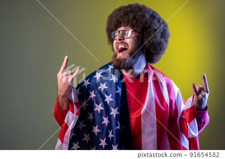 Excited happy hipster man with Afro hairstyle wrapped in american flag, rejoicing, celebrating national holiday, showing v sign. Indoor studio shot isolated on colorful neon light background. Excited happy hipster man with Afro hairstyle wrapped in american flag, rejoicing, celebrating national holiday, showing v sign. Indoor studio shot isolated on colorful neon light background. 91654582