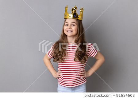 Portrait of satisfied arrogance little girl princess wearing striped T-shirt and golden diadem crown, looking at camera keeps hands on hips. Indoor studio shot isolated on gray background. Portrait of satisfied arrogance little girl princess wearing striped T-shirt and golden diadem crown, looking at camera keeps hands on hips. Indoor studio shot isolated on gray background. 91654608