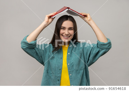 Funny positive woman holding opened book on head, student making ritual to remember lot of study material, wearing casual style jacket. Indoor studio shot isolated on gray background. Funny positive woman holding opened book on head, student making ritual to remember lot of study material, wearing casual style jacket. Indoor studio shot isolated on gray background. 91654618