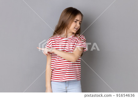 Portrait of dark haired little girl wearing striped T-shirt showing exit, demanding to leave her alone, has resentful irritated expression. Indoor studio shot isolated on gray background. 91654636