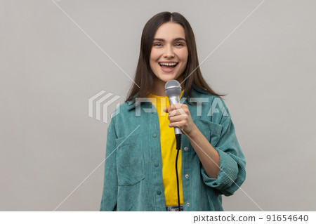Portrait of positive woman reporter standing with microphone in hands, sing songs with happy expression or telling news, wearing casual style jacket. Indoor studio shot isolated on gray background. 91654640