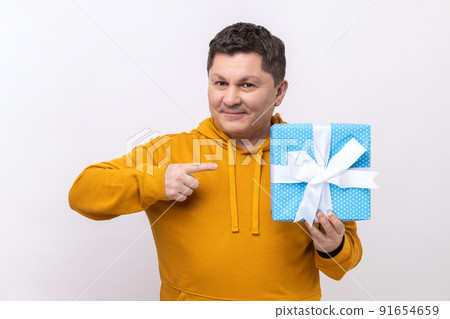 Delighted middle aged man with dark hair holding blue gift box with smile, pointing finger, looking at camera, wearing urban style hoodie. Indoor studio shot isolated on white background. 91654659