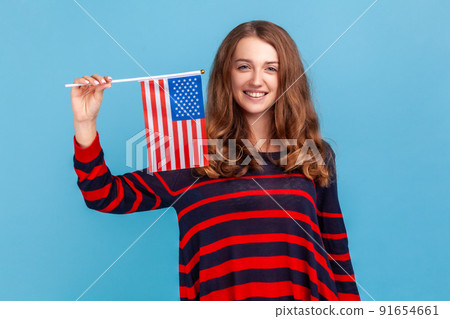 Good looking woman wearing striped casual style sweater, waving united states of america flag, celebrating national holiday, patriotism, independence. Indoor studio shot isolated on blue background. 91654661