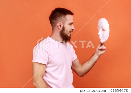 Side view portrait of bearded man looking at white mask in hands with attentive look, trying to understand hiding personality, wearing pink T-shirt. Indoor studio shot isolated on orange background. 91654678