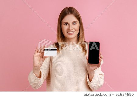 Portrait of smiling optimistic blond woman holding credit card and cell phone with black black screen, online banking, wearing white sweater. Indoor studio shot isolated on pink background. Portrait of smiling optimistic blond woman holding credit card and cell phone with black black screen, online banking, wearing white sweater. Indoor studio shot isolated on pink background. 91654690