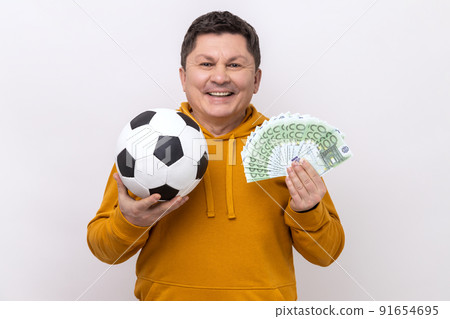 Smiling middle aged man showing soccer ball and fun of hundred euro bills, winning lot of money betting for sport, wearing urban style hoodie. Indoor studio shot isolated on white background. 91654695