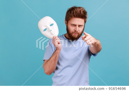 Portrait of strict aggressive young adult bearded man holding white mask in hand, pointing finger to camera with serious bossy expression. Indoor studio shot isolated on blue background. 91654696