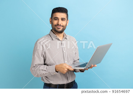 Smiling man freelancer with beard standing and holding laptop, satisfied with teleworking, likes his job, looking at camera, wearing striped shirt. Indoor studio shot isolated on blue background. 91654697