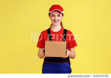 Portrait of delighted courier woman standing with cardboard parcel, giving it to client, looking at camera, wearing overalls and red cap. Indoor studio shot isolated on yellow background. Portrait of delighted courier woman standing with cardboard parcel, giving it to client, looking at camera, wearing overalls and red cap. Indoor studio shot isolated on yellow background. 91654750