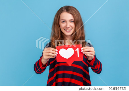 Portrait of optimistic woman wearing striped casual style sweater, holding heart like icon of social media and looking at camera with toothy smile. Indoor studio shot isolated on blue background. Portrait of optimistic woman wearing striped casual style sweater, holding heart like icon of social media and looking at camera with toothy smile. Indoor studio shot isolated on blue background. 91654785