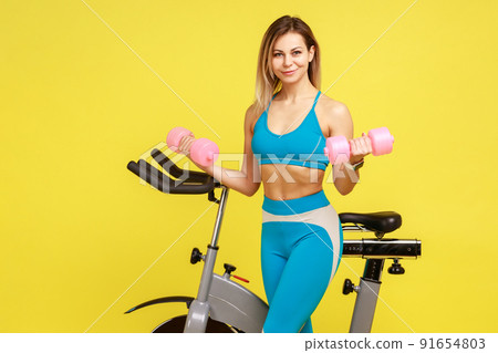 Portrait of beautiful blonde woman fitness instructor doing exercises with dumbbells for biceps after cardio workout, wearing blue sportswear. Indoor studio shot isolated on yellow background. 91654803