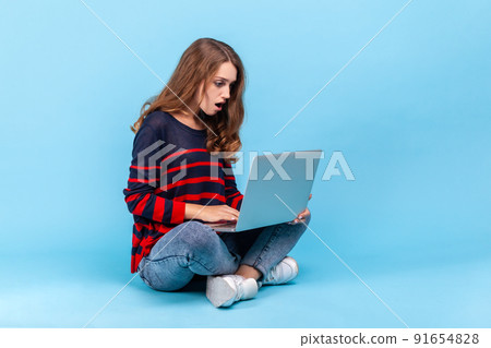 Astonished woman wearing striped casual style sweater, sitting on floor with crossed legs and looking at laptop screen with shocked expression. Indoor studio shot isolated on blue background. Astonished woman wearing striped casual style sweater, sitting on floor with crossed legs and looking at laptop screen with shocked expression. Indoor studio shot isolated on blue background. 91654828
