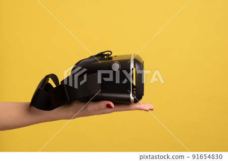Side view of woman hand holding VR goggles, virtual reality 3d glasses headset for cyberspace simulation, innovative technologies, future and science. Indoor studio shot isolated on yellow background. 91654830
