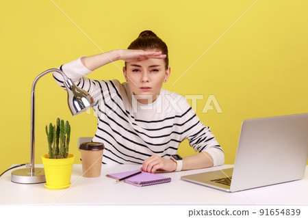 Portrait of young woman employee sitting at workplace with laptop and looking far away with hand above eyes, searching with view. Indoor studio studio shot isolated on yellow background. Portrait of young woman employee sitting at workplace with laptop and looking far away with hand above eyes, searching with view. Indoor studio studio shot isolated on yellow background. 91654839
