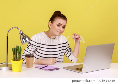 Portrait of disappointed upset woman manager sitting at workplace with laptop and showing a little bit gesture, measuring minimum. Indoor studio studio shot isolated on yellow background. Portrait of disappointed upset woman manager sitting at workplace with laptop and showing a little bit gesture, measuring minimum. Indoor studio studio shot isolated on yellow background. 91654846