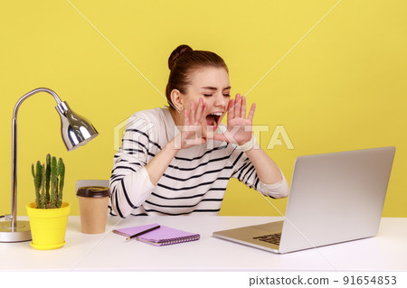 Nervous angry woman office worker in striped shirt loudly screaming holding hands near mouth, talking video conference sitting at workplace. Indoor studio studio shot isolated on yellow background. 91654853