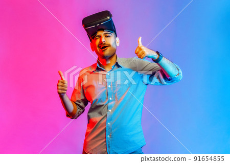 Portrait of man in shirt with virtual reality glasses expressing excitement after playing game simulator, standing with thumbs up. Indoor studio shot isolated on colorful neon light background. 91654855