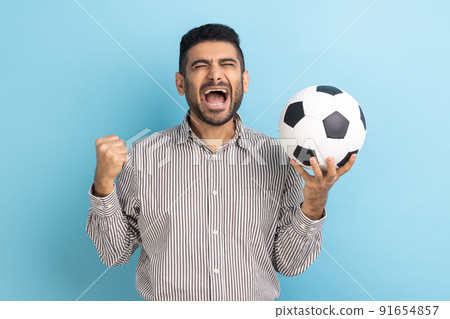 Bearded businessman screaming widely opening mouth, celebrating victory of favourite football team, holding soccer ball in hands, wearing striped shirt. Indoor studio shot isolated on blue background. 91654857