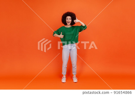 Full length portrait of happy friendly woman with Afro hairstyle wearing green casual style sweater standing with thumbs up, expressing positive. Indoor studio shot isolated on orange background. Full length portrait of happy friendly woman with Afro hairstyle wearing green casual style sweater standing with thumbs up, expressing positive. Indoor studio shot isolated on orange background. 91654866