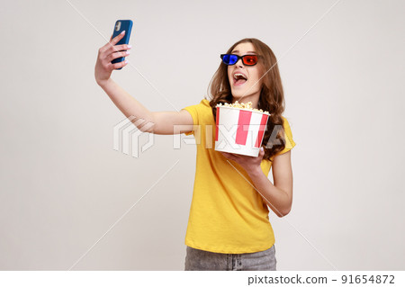 Young funny happy excited teenager girl wearing yellow casual style T-shirt holding takeaway popcorn bucket, making selfie or broadcasting livestream. Indoor studio shot isolated on gray background. Young funny happy excited teenager girl wearing yellow casual style T-shirt holding takeaway popcorn bucket, making selfie or broadcasting livestream. Indoor studio shot isolated on gray background. 91654872