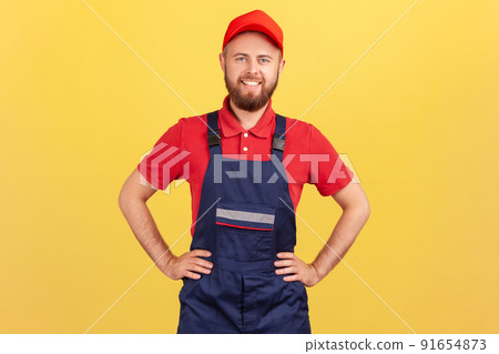 Portrait of confident worker man standing with hands on hips, looking at camera, being ready to take over and complete order, wearing overalls and cap. Indoor studio shot isolated on yellow background 91654873