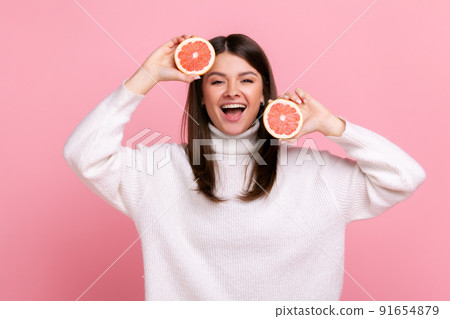 Excited woman with slices of grapefruit in hands, looking at camera, expressing positive emotions, wearing white casual style sweater. Indoor studio shot isolated on pink background. Excited woman with slices of grapefruit in hands, looking at camera, expressing positive emotions, wearing white casual style sweater. Indoor studio shot isolated on pink background. 91654879