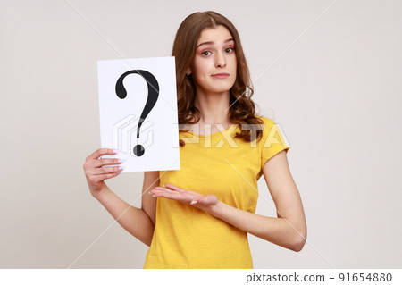 Young attractive brown haired young woman in yellow casual style T-shirt holding paper with question mark, making decision, solving problem. Indoor studio shot isolated on gray background. 91654880