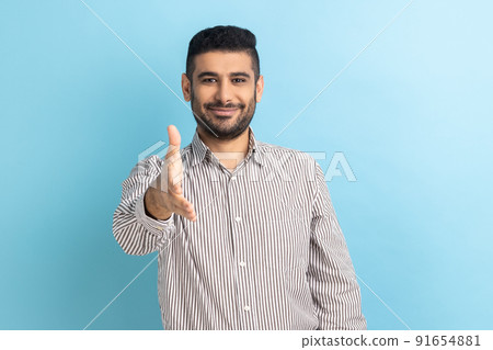 Portrait of handsome happy businessman standing and looking at camera with toothy smile and giving hand to greeting or handshake, wearing striped shirt. Indoor studio shot isolated on blue background. Portrait of handsome happy businessman standing and looking at camera with toothy smile and giving hand to greeting or handshake, wearing striped shirt. Indoor studio shot isolated on blue background. 91654881