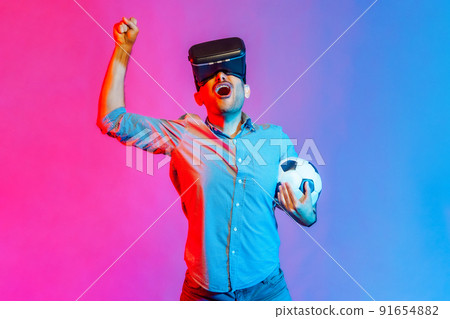 Portrait of man in shirt wearing headset and holding soccer ball, watching 3d football team, cheering, celebrating victory. Indoor studio shot isolated on colorful neon light background. 91654882