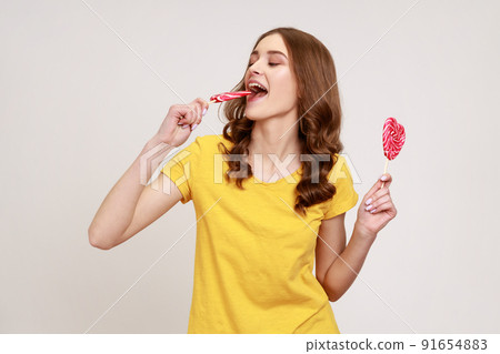 Portrait of woman in yellow casual T-shirt biting lollipop, tasting sweet heart shape candy with satisfied expression, enjoying delicious flavor dessert. Indoor studio shot isolated on gray background 91654883