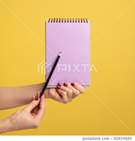 Profile side view closeup of woman hand holding purple notepad in hand and showing empty paper with pen or pencil. Indoor studio shot isolated on yellow background. 91654884