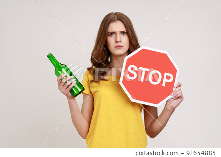 Stop drinking alcohol. Serious teenager girl in yellow T-shirt holding red stop sign and alcohol bottle, looking at camera strict expression. Indoor studio shot isolated on gray background. 91654885