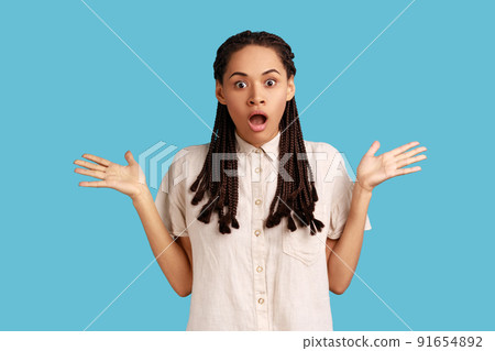 Indignant shocked woman with black dreadlocks has surprised angry expression, raised arms, looking at camera with big eyes, wearing white shirt. Indoor studio shot isolated on blue background. 91654892
