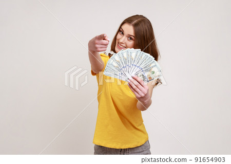 Portrait of positive young woman holding fan of hundred dollar bills and pointing finger at camera, choosing you as winner of money jack pot. Indoor studio shot isolated on gray background. 91654903