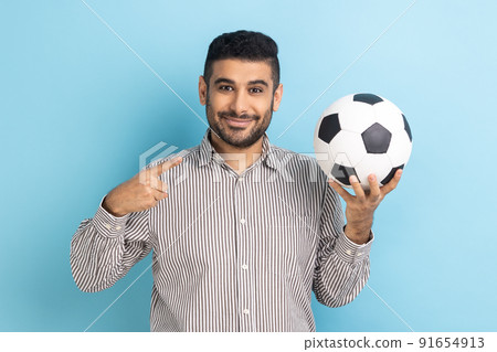 Portrait of handsome businessman pointing finger at soccer ball on his hand with smiling positive expression, wearing striped shirt. Indoor studio shot isolated on blue background. Portrait of handsome businessman pointing finger at soccer ball on his hand with smiling positive expression, wearing striped shirt. Indoor studio shot isolated on blue background. 91654913