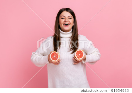 Happy optimistic playful girl holding two half slices of grapefruit in front of her breast, laughing, wearing white casual style sweater. Indoor studio shot isolated on pink background. 91654923