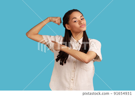 Self assured woman with dreadlocks with pleasant appearance, raises hand and indicates at biceps, confirms women can be also strong, wearing white shirt. Indoor studio shot isolated on blue background 91654931