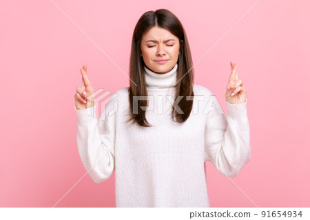 Pretty woman standing with closed eyes and crossing fingers, wishing for good luck, waiting miracle, wearing white casual style sweater. Indoor studio shot isolated on pink background. Pretty woman standing with closed eyes and crossing fingers, wishing for good luck, waiting miracle, wearing white casual style sweater. Indoor studio shot isolated on pink background. 91654934