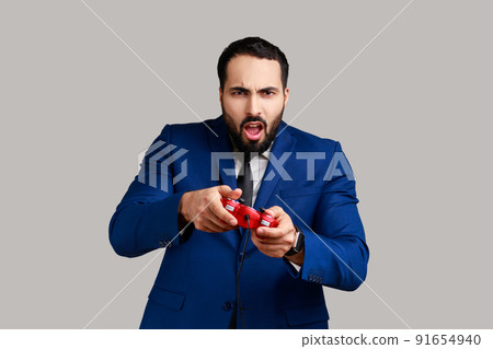Concentrated attractive bearded man holding in hands red gamepad joystick, grimacing playing video games, wearing official style suit. Indoor studio shot isolated on gray background. Concentrated attractive bearded man holding in hands red gamepad joystick, grimacing playing video games, wearing official style suit. Indoor studio shot isolated on gray background. 91654940