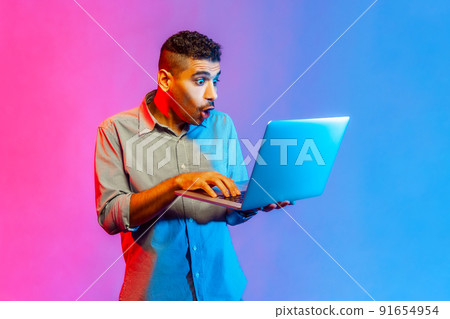 Portrait of man in shirt doing freelance job on laptop, typing email or surfing internet, looking surprised at laptop screen. Indoor studio shot isolated on colorful neon light background. 91654954