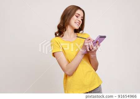 Portrait of glad curly haired teenage girl in yellow casual style T-shirt writing in notebook, filling diary or schedule, smiling sincerely. Indoor studio shot isolated on gray background. 91654966