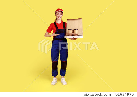 Full length portrait of courier woman presenting coffee and boxes with pizza, offering drinks and food delivery, wearing overalls and red cap. Indoor studio shot isolated on yellow background. Full length portrait of courier woman presenting coffee and boxes with pizza, offering drinks and food delivery, wearing overalls and red cap. Indoor studio shot isolated on yellow background. 91654979