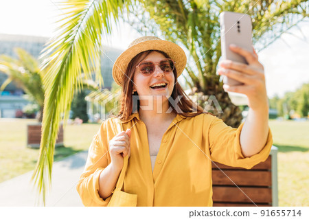 Happy redhead woman in straw hat and sunglasses take a selfie at vacation on a tropical island, palm trees in the background. Solo Female Traveler. Summer concept. 91655714