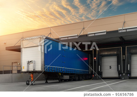Innovative modern logistics warehouse center complex building exterior bay gate semi-trailer unloading goods distribuition warm blue sunset sky background. Cargo storage facility industry building 91656786