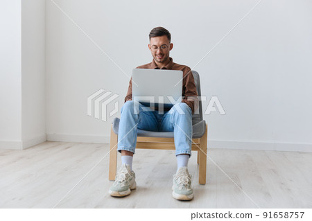 Smiling happy young tanned man in eyewear hold laptop on knees chatting with friends typing message looks at screen sitting on chair at white home wall. Lottery winner offer. Copy space Smiling happy young tanned man in eyewear hold laptop on knees chatting with friends typing message looks at screen sitting on chair at white home wall. Lottery winner offer. Copy space 91658757