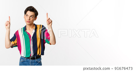 Attractive gay man looking thoughtful, pointing fingers up at logo, standing over white background Attractive gay man looking thoughtful, pointing fingers up at logo, standing over white background 91676473