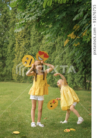 Two amusing girls with a variety of cardboard emoticons in their hands are having fun in the park on the lawn. Children indulge and make faces 91676755