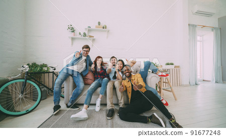 Young friends sitting on sofa smiling and showing thumbs up all together at home indoors 91677248