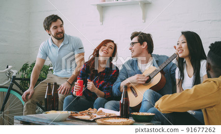 Happy young friends have party at shared apartment and singing together while their friend playing guitar at home indoors 91677262